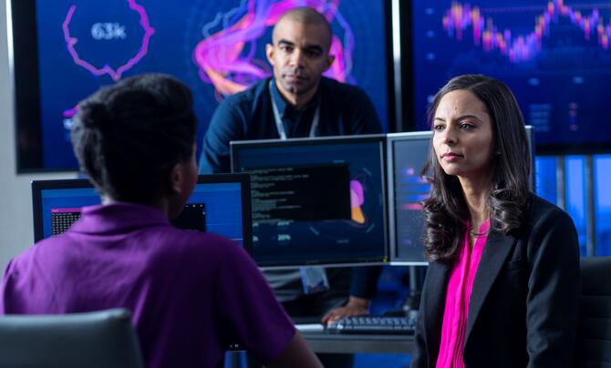 Three employees discussing something with charts on a screen behind them