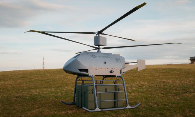 A small, helicopter-like uncrewed aerial system landed on a grassy field over an Army-green container.
