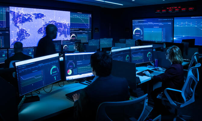 Command center with multiple people and multiple computer and projector screens showing data, charts and maps.