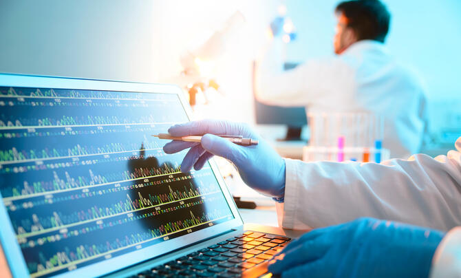 A gloved hand with a white coat points to a laptop screen displaying unspecified data, with a white-coated man in the background.