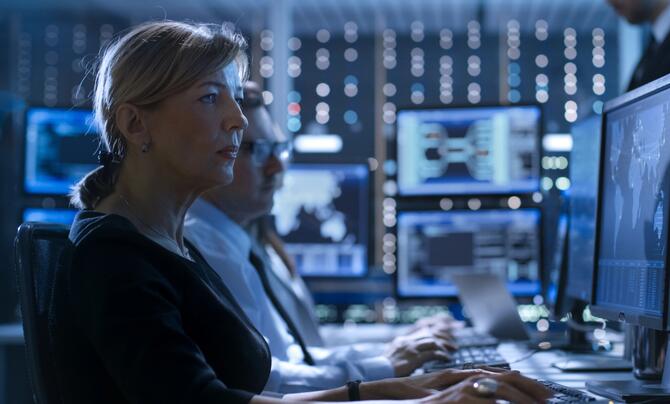 A woman and a man seated in a data center with multiple computer monitors. 