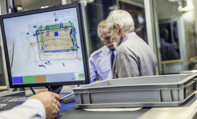 In foreground is computer monitor with luggage image, security guard and senior man in background.