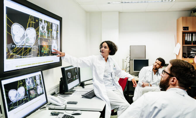 A clinical registrar reviewing a patients test results, looking at some information on a large monitor with some other doctors.