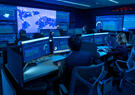 Analysts monitor data dashboards and a global map across multiple screens within a dimly lit, blue-toned security operations center.