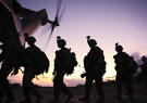 Silhouetted soldiers carrying gear walk toward a helicopter at sunset, its rotor blades spinning overhead. The scene is backlit by a warm sky, emphasizing their movement and mission readiness.