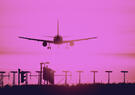 Airplane descends for landing against a vivid pink sky, silhouetted above runway lights and approach equipment.
