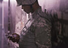 A soldier in camouflage uniform and helmet leans against a wall, looking down at a smartphone. The scene is softly lit with a muted, slightly somber tone.