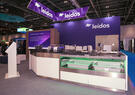 A trade show booth with prominent “Leidos” branding features a modern display counter and seating area. Overhead signage and screens highlight the company’s presence in a large exhibition hall.