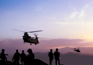 Six soldiers in silhouette standing on top of a large hill or mountain facing the sunset while a Chinook and other unidentified helicopter fly past.