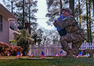 A male soldier bends on one knee to hug a child in front of a home along a path with green grass and small U.S. flags along the path.