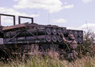 A soldier in tactical gear inspects a truck-mounted system loaded with cylindrical equipment in a grassy field under a partly cloudy sky.