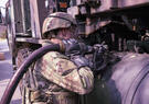 A soldier in protective gear refuels a military vehicle using a large fuel hose and heavy-duty equipment.