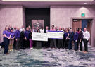 A group of employees stands together indoors holding oversized donation checks for Food Bank of the Rockies during a community fundraising event.