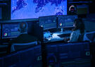 Two analysts monitor cybersecurity dashboards and global network activity on large screens in a high-tech operations center.