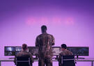 Military personnel monitor surveillance and operational data on multiple screens in a command center illuminated by purple lighting.