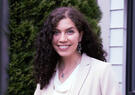 A smiling woman with curly dark hair stands outdoors wearing a light-colored blazer and earrings, with greenery in the background.