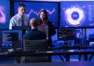 Three professionals collaborate in a cybersecurity operations center, reviewing live dashboards and data visualizations on multiple monitors to monitor network activity and respond to potential threats.