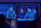 A team of professionals collaborates around a digital workspace, reviewing data and plans in a technology operations center illuminated by large interactive displays.