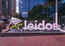 A large Leidos logo sign stands outside a modern glass office building, surrounded by landscaped greenery and brick walkways.