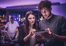 Two professionals collaborate over a tablet in a technology-focused workspace, reviewing information together under ambient purple lighting.