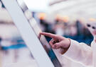 Close-up of a traveler using a touchscreen self-service kiosk while holding a smartphone, illustrating digital identity verification and contactless passenger processing at an airport.