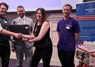 Leidos employees present a donated laptop to representatives of the Jon Egging Trust during a community partnership event, standing beside a Jon Egging Trust display banner.