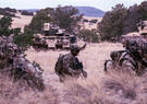 Soldiers move through rugged terrain during a military training exercise, with armored combat vehicles providing support in the background.