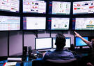Two professionals monitor operational dashboards on multiple large displays, reviewing performance metrics, analytics, and system status in a technology operations center.