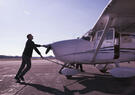 A person pushes a small single-engine aircraft across an airport ramp before flight, with another individual boarding through the open cabin door under a clear sky.