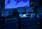 A dark operations control command center, with two people sitting at desks viewing computer screens.