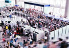Crowds of travelers move through security screening checkpoints and queues inside a busy airport terminal.
