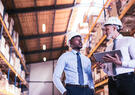 Two professionals wearing hard hats discuss operations while reviewing information on a tablet in a warehouse.