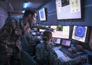 Two military servicemembers in a small computer operations room viewing and pointing at computer screens showing simulated data and maps 