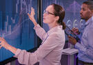 Two professionals analyze data visualizations on large digital displays, with one pointing to a chart while the other holds a tablet.
