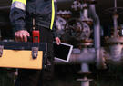 Industrial worker in high-visibility clothing carries a toolbox and tablet beside large pipes and valves at an industrial facility.