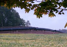 Large black gas pipeline sections stretch across a grassy field bordered by trees, with wildflowers in the foreground.