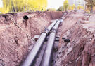 Two large metal pipelines run through an excavated trench at a construction site, surrounded by earth and construction materials.