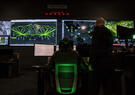 Two people facing away looking at large computer and TV screens with various data feeds in a dark room