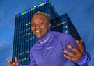Raven Saunders posing in a Leidos pullover in front of Leidos Global Headquarters. The building is lit up in green and purple.