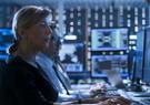 A woman and a man seated in a data center with multiple computer monitors. 