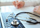 Stethoscope laying on a laptop keyboard with a person in a white coat writing in the background.