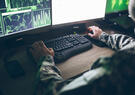 A soldier sits at a desk with a computer monitor and keyboard