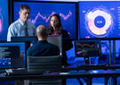 Two men and a women in a computer lab with large screens showing data readouts