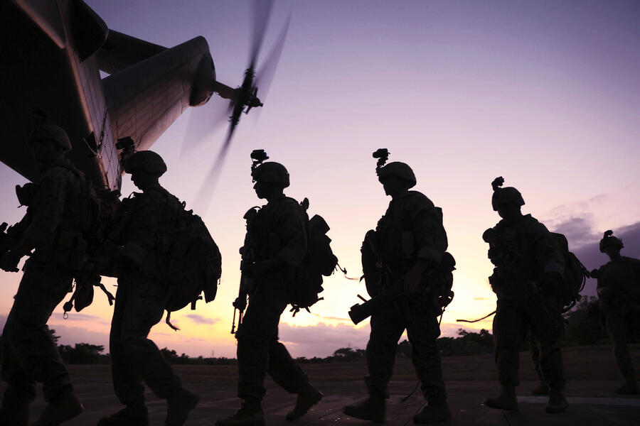 Silhouetted soldiers carrying gear walk toward a helicopter at sunset, its rotor blades spinning overhead. The scene is backlit by a warm sky, emphasizing their movement and mission readiness.