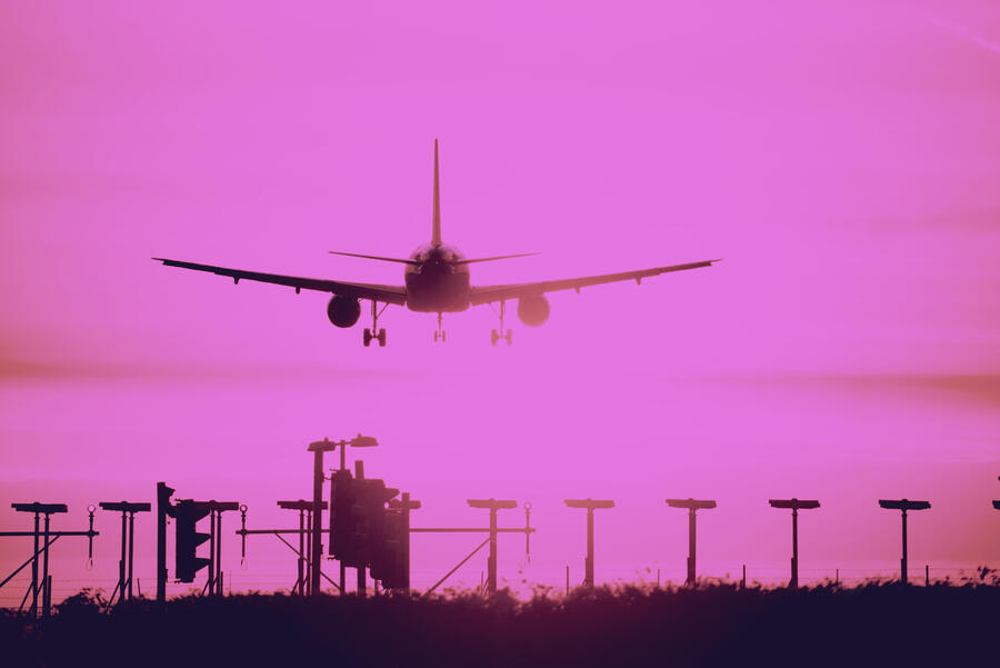 Airplane descends for landing against a vivid pink sky, silhouetted above runway lights and approach equipment.