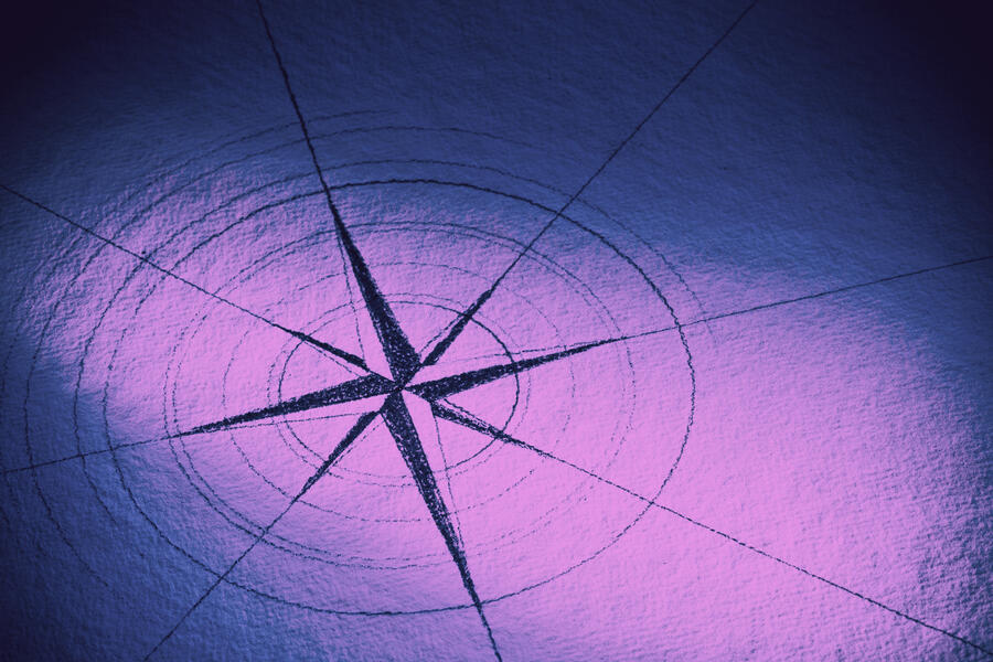 A stylized compass rose is drawn on a textured surface, illuminated by purple and blue lighting. The design features radiating lines and circular markings centered on a star-like shape.