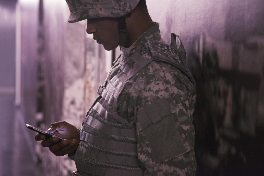 A soldier in camouflage uniform and helmet leans against a wall, looking down at a smartphone. The scene is softly lit with a muted, slightly somber tone.