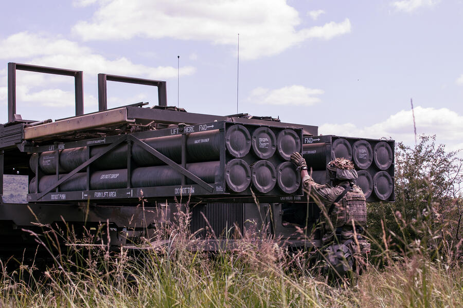 A soldier in tactical gear inspects a truck-mounted system loaded with cylindrical equipment in a grassy field under a partly cloudy sky.