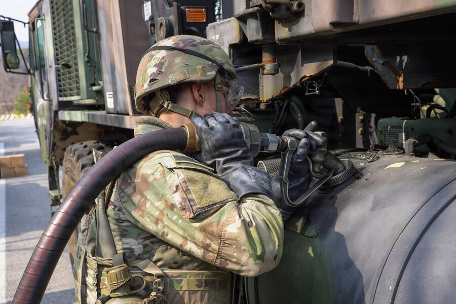 A soldier in protective gear refuels a military vehicle using a large fuel hose and heavy-duty equipment.