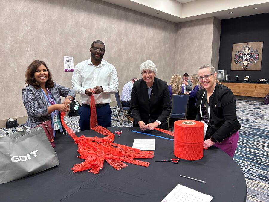A group of employees smiles while assembling orange mesh produce bags around a table during a volunteer community service event.